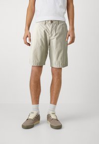 Shorts en coton beige clair avec une taille élastique, dotés d'une fermeture à bouton unique et de poches latérales, associés à des chaussures grises et des chaussettes blanches.