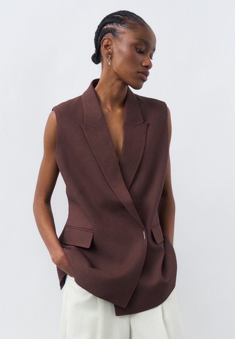 Γυναίκα με πλεξούδες στα μαλλιά, που φορά αμάνικο καφέ σακάκι και άσπρο παντελόνι, στέκεται με τα χέρια στις τσέπες, κοιτάζοντας προς τα κάτω πλάγια.