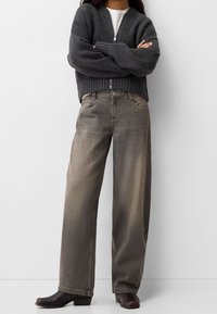 Grå zip-up stickad tröja med ribbad textur, tillsammans med vida beige jeans med en lätt gradient. Svarta klackade ankelskor.