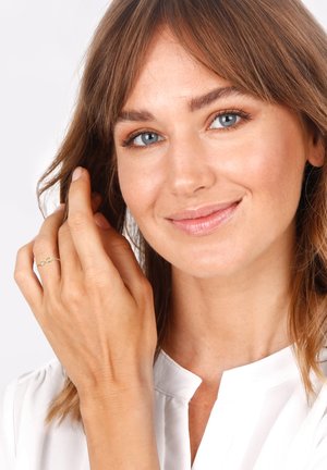 Goldring mit einem Unendlichkeitssymbol, getragen an einer Hand, die braunes Haar berührt. Das Model hat blaue Augen, trägt eine weiße Bluse vor einem neutralen Hintergrund.
