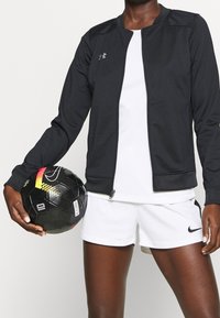 Black athletic zip-up jacket with gray accents, worn over a white shirt and shorts. The model holds a black and yellow soccer ball.