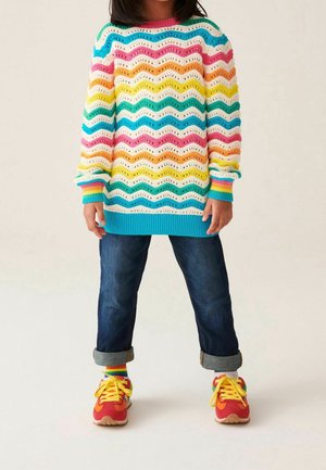 Pull en tricot multicolore avec un motif en vagues, ourlet côtelé bleu, associé à un jean en denim retroussé et des baskets rouges avec des lacets jaunes.