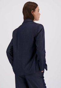 Mörkblå skräddarsydd kavaj med en strukturerad design, enkel slits och långa ärmar; tillverkad av ett slätt, texturerat tyg.