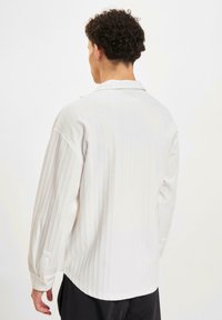 Chemise blanche à manches longues avec des rayures verticales, un col, un poignet boutonné et une coupe décontractée, mettant en valeur une texture de tissu lisse.