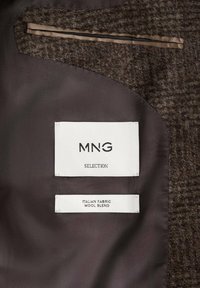 Tessuto in misto lana di colore marrone con finitura testurizzata. Le etichette bianche riportano la scritta "MNG SELECTION" e "ITALIAN FABRIC WOOL BLEND", cucite all'interno del capo.