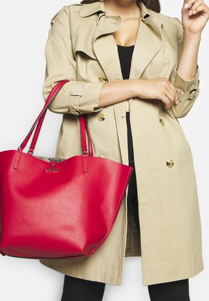 Kvinde iført beige trenchcoat og sort tøj, der holder en stor rød skuldertaske på venstre arm.