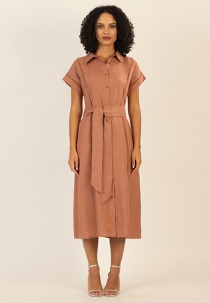 Woman with curly hair wears a buttoned, short-sleeve, midi-length brown dress with a tied waist belt and silver high-heeled sandals.