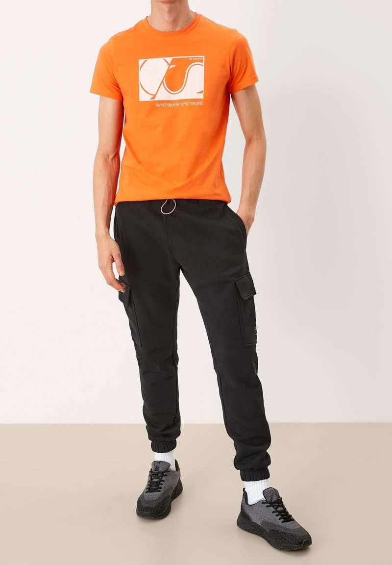 Orange t-shirt med grafiskt tryck över svarta cargobyxor. Byxorna har sidofickor, ribbade muddar, och är parat med grå sneakers och vita strumpor.
