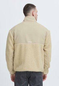 Giacca in pile beige con una texture morbida, tessuto liscio a contrasto sulle spalle, colletto alto e una piccola etichetta del marchio sulla parte posteriore.