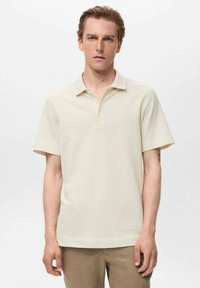 Cămașă polo bej cu mânecă scurtă, dintr-un material texturat, cu guler butonat și tiv drept. Pur’tă cu pantaloni maro deschis, modelul stând în fața unui fundal simplu.