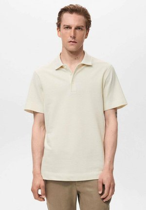Cămașă polo bej cu mânecă scurtă, dintr-un material texturat, cu guler butonat și tiv drept. Pur’tă cu pantaloni maro deschis, modelul stând în fața unui fundal simplu.