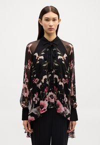 Blouse noire à motifs floraux avec de longues manches transparentes, motifs floraux en rose et vert, nœud au cou, ourlet irrégulier, tissu léger.