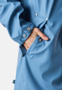 Giacca impermeabile azzurro chiaro con finitura testurizzata, dotata di bottoni a pressione e una tasca laterale. Include un piccolo dettaglio a bandiera sull'orlo.