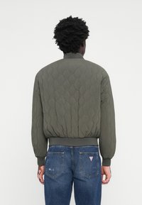 Tömbösített zöld dzseki kerek gallérral, rugalmas mandzsettákkal és derékrésszel. Kék farmernadrággal párosítva, amely texturált mintáját mutatja.