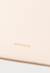 Világos rózsaszín műbőr táska texturált felülettel. Az alján aranyozott, dombornyomott "ANNA FIELD" logó látható. Egyéb részletek nem láthatók.