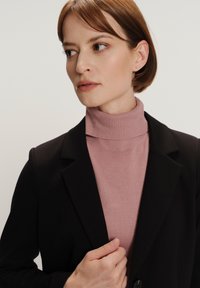 Zwarte blazer met notch-lapels over een lichtroze coltrui. De blazer heeft een gladde textuur, terwijl de coltrui een geribbelde afwerking heeft.