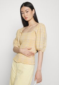 Blusa amarela xadrez com mangas bufantes, corpete franzido e decote quadrado. Combinada com calças de corduroy amarelas claras.