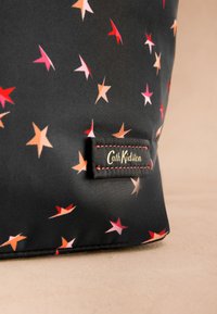 Juoda medžiaga su rožinių, raudonų ir oranžinių žvaigždžių raštu bei Cath Kidston etiketė su raudonais siūlais ant smėlio spalvos paviršiaus.