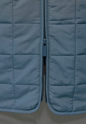 Vadderad blå väst med en slät finish. Har en central dragkedja med en gummihandtag och ett rutmönster över tyget.