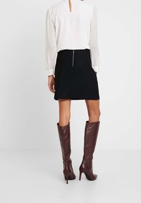 Blusa bianca con dettagli a chiave sul retro, abbinata a una mini gonna nera con zip sul retro. Stivali alti fino al ginocchio marroni con un design aderente.