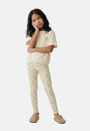 Jeune fille aux longs cheveux foncés portant une chemise beige avec broderie florale, des leggings fleuris et des chaussures beiges sans lacets, debout la main sur la joue.