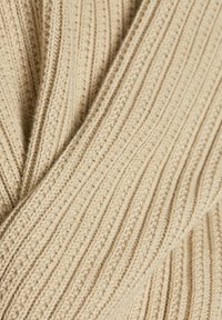 Beige gebreide stof met een geribbelde textuur en verticale lijnen, versierd met kleine contrasterende stippen die diepte en interesse toevoegen.