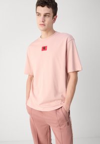 T-shirt en coton rose clair avec un patch logo rouge carré sur la poitrine. Col rond, manches courtes et coupe décontractée. Associé à un pantalon assorti.
