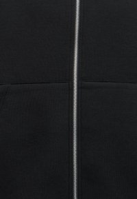 Tissu noir avec une fermeture éclair métallique argentée qui traverse verticalement le centre, et deux lignes de couture horizontales près du milieu.
