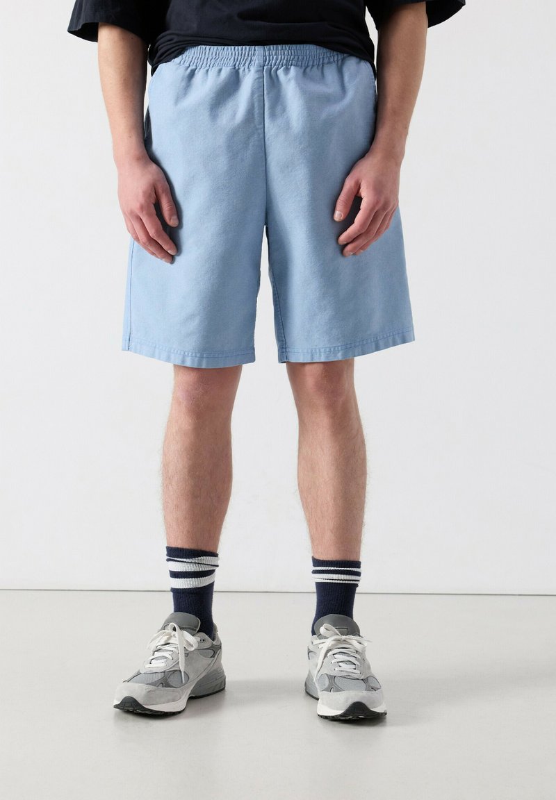 Mees, kes kannab helesiniseid elastse vöökoha jaotusega lühikesi pükse, tumesiniseid triibulisi sokke, halli spordijalatseid ja musta särki, käed mõlemal pool rippumas.