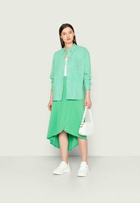 Camisa de botão verde e branca com riscas, sobre uma saia assimétrica verde sólida, combinada com ténis brancos e uma pequena mala branca.