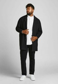 Schwarzer langer Mantel über einem weißen Hemd mit Knöpfen, kombiniert mit schwarzen Hosen und weißen Sneakers. Der Mantel hat ein einfaches Design und zwei vordere Knöpfe.
