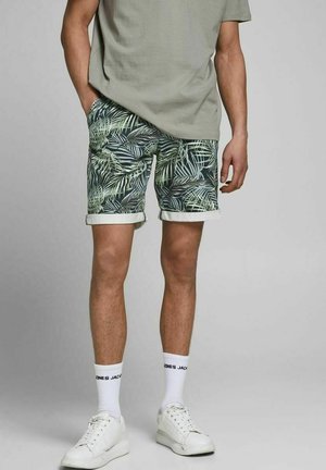 Homme portant un short vert à imprimé feuilles tropicales, un t-shirt gris clair, des chaussettes blanches et des baskets blanches, debout sur un fond gris.