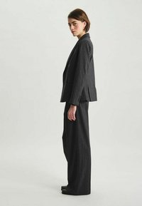 Costume noir à rayures, coupe ajustée. Blazer à double boutonnage avec revers crantés, pantalons à jambes larges, texture lisse et rayures contrastées.