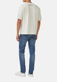 Mann trägt ein weißes T-Shirt mit kurzen Ärmeln und Text auf dem oberen Rücken, blaue Jeans und weiße Turnschuhe, steht mit dem Rücken zur Kamera vor einem weißen Hintergrund.