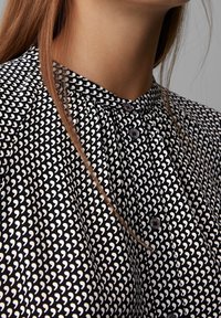 Svart blus med ett vitt hjärtmönster, plisserad halsringning och runda knappar. Tyget har en mjuk textur.