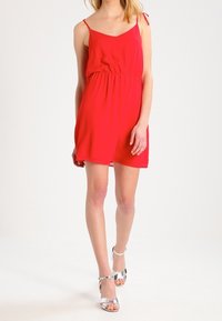 Rotes ärmelloses Kleid mit tailliertem Schnitt, leichtem Stoff, knielang, ausgestattet mit dünnen Trägern. Kombiniert mit silbernen Riemchensandalen.