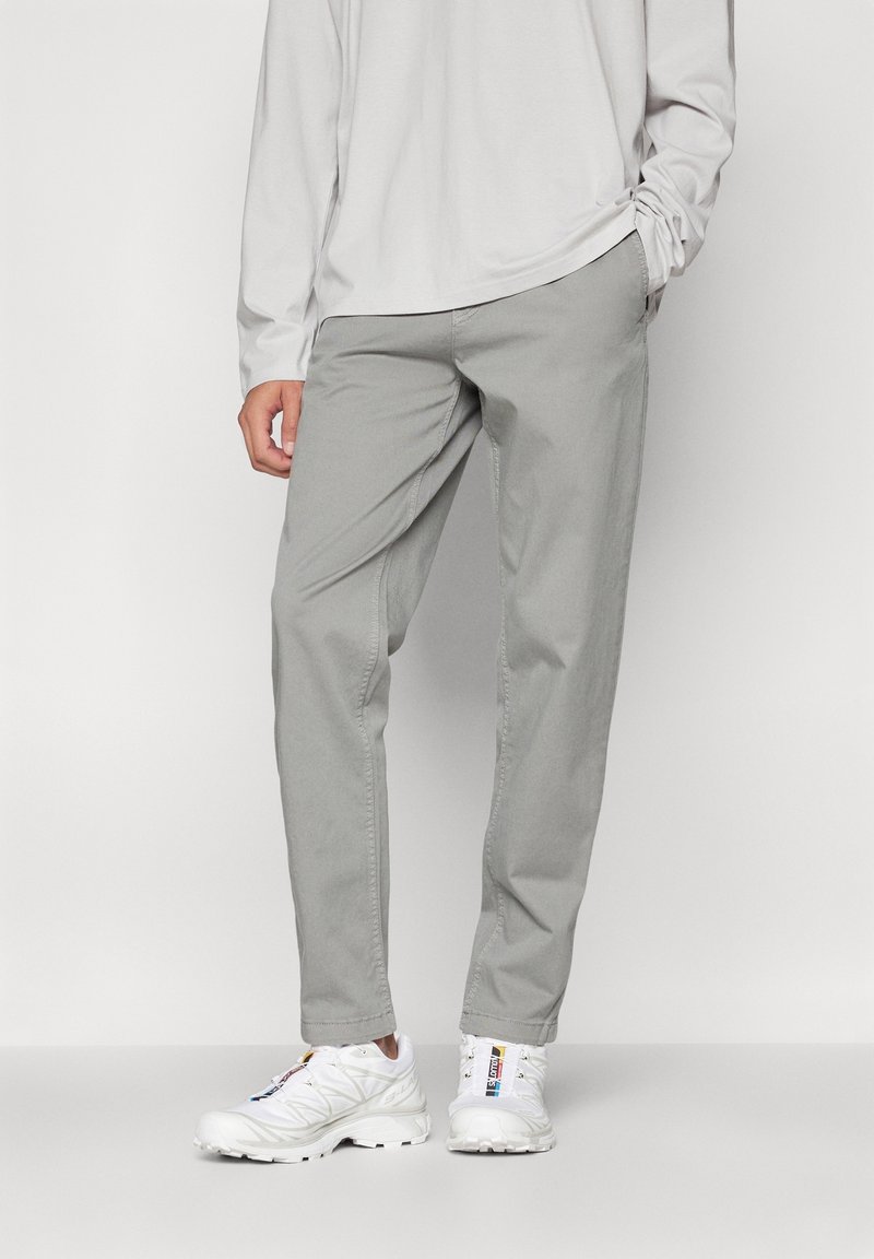 Personne portant un pantalon décontracté gris clair, une chemise blanche à manches longues et des baskets blanches avec des languettes colorées, debout devant un fond uni.