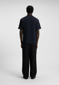 Muž s krátkými kudrnatými vlasy, oblečený v námořnickém modrém tričku s krátkým rukávem a volných černých kalhotách, stojící zády k nám na jednobarevném světlém pozadí.