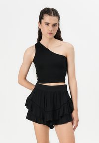 Svart crop top med én skulder laget av ribbet stoff, kombinert med lagdelte shorts med volanger i svart, som har en fast linning og dekorative detaljer.