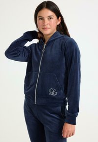 Hoodie zippé en velours bleu marine avec une poche avant et un logo brodé. Texture douce, poignets côtelés et une capuche pour un confort supplémentaire.