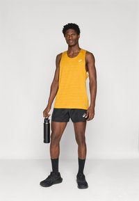 Κίτρινο tank top με διακριτική υφή και μαύρο λογότυπο, συνδυασμένο με μαύρες βερμούδες, μαύρες κάλτσες και αθλητικά παπούτσια, κρατώντας ένα σκούρο μπουκάλι νερού.