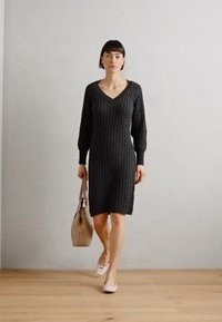 Tamsiai pilkas raukšlėtas megztinis su V formos iškirpte, ilgomis rankovėmis ir prigludusia siluetu. Modelis laiko šviesiai smėlinį rankinuką, stovėdamas ant šviesių medinių grindų.