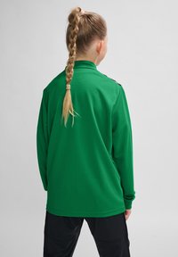 Lange mouwen, groene sporttop van een textuurstof, met een opstaande kraag en opvallende schouderaccenten. Van de achterkant bekeken.