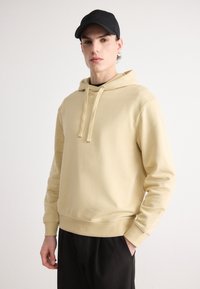Felpa beige con cappuccio, dotata di tasca a marsupio e cordino, realizzata in cotone, con polsini e orlo a costine. Cappellino nero indossato.