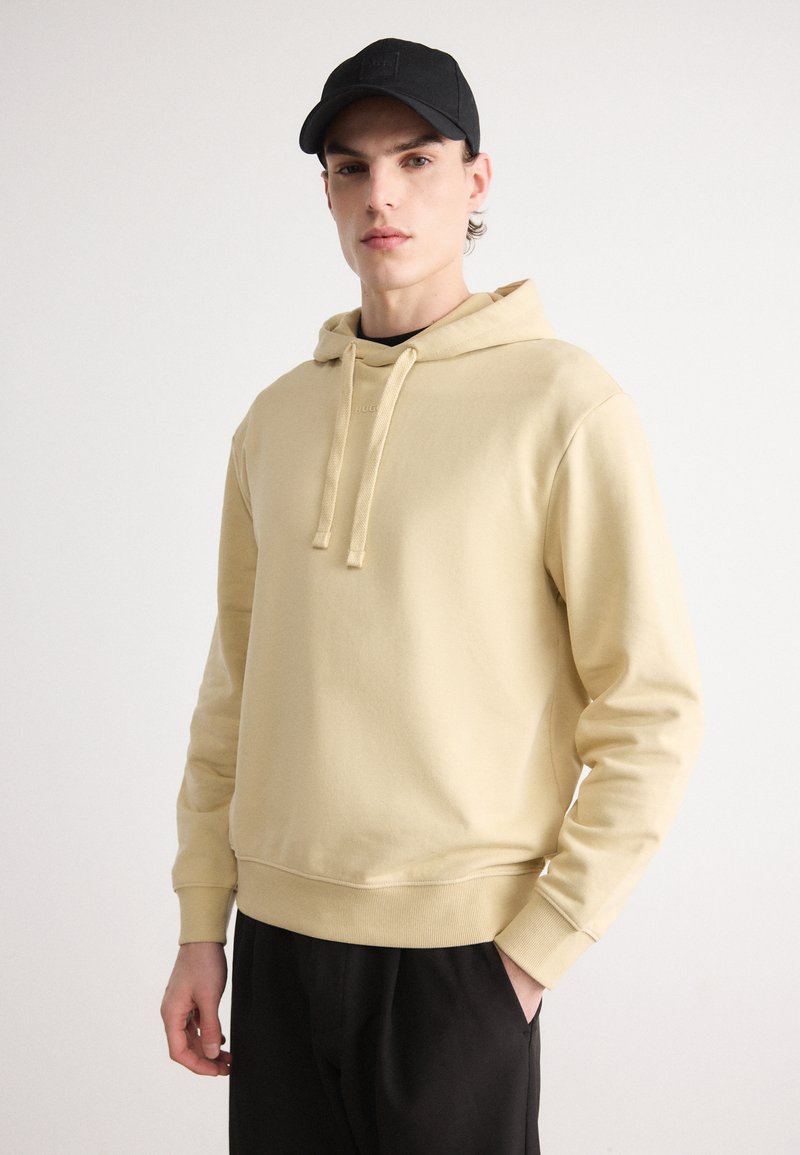 Felpa beige con cappuccio, dotata di tasca a marsupio e cordino, realizzata in cotone, con polsini e orlo a costine. Cappellino nero indossato.