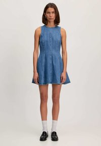 Denim jurk met mouwloos ontwerp, aansluitende lijfje, flared rok, lichtblauwe kleur, zichtbare stiksels en ruwe zoom. G gedragen met zwarte schoenen en witte sokken.
