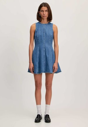 Denim jurk met mouwloos ontwerp, aansluitende lijfje, flared rok, lichtblauwe kleur, zichtbare stiksels en ruwe zoom. G gedragen met zwarte schoenen en witte sokken.