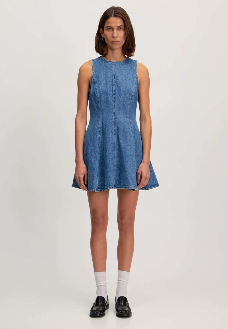 Denim jurk met mouwloos ontwerp, aansluitende lijfje, flared rok, lichtblauwe kleur, zichtbare stiksels en ruwe zoom. G gedragen met zwarte schoenen en witte sokken.