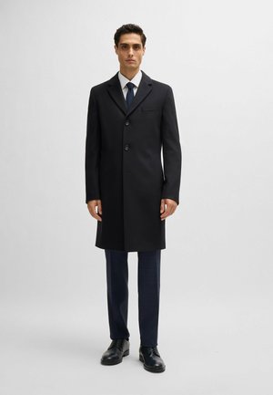 Manteau en laine noir avec un design à un seul bouton, trois boutons et une poche poitrine. Le modèle porte un pantalon habillé bleu marine et des chaussures noires.