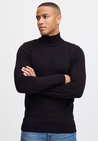 Suéter negro de cuello alto hecho de tela suave, con mangas largas y puños y dobladillo acanalados. Lleva puesto unos jeans de mezclilla azul claro.